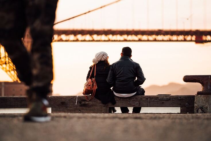 A couple with hearing loss is dressed for cold weather and sitting on a dock by a bridge.