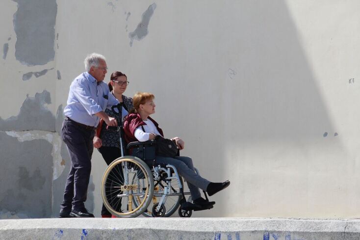 A woman with an ear stroke and hearing loss sits in a wheelchair, pushed by a man and accompanied by a nurse.