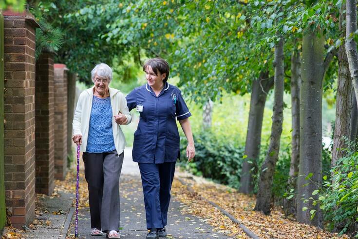 A nurse helps a woman holding a purple cane walk on a pathway between a brick wall and a row of trees with fallen leaves.