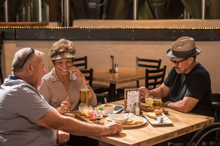 A man socializes at a restaurant table with a woman in a visor and a man with a hat and glasses sitting in a wheelchair.