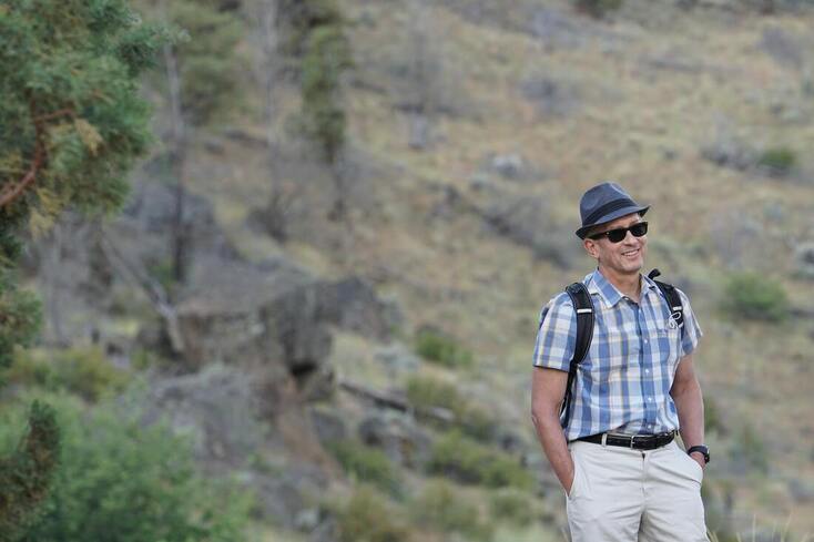 A backpacker in khakis and a plaid shirt wearing a grey-blue fedora hat and sunglasses smiles with his hands in his pocket.