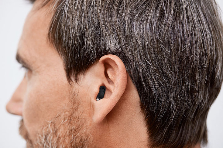 A man with salt-and-pepper hair and a beard wears a black completely-in-the-canal hearing aid.