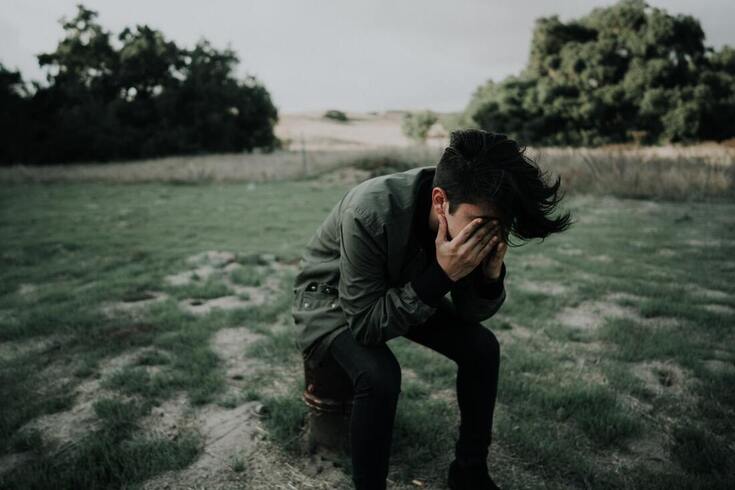 man with tinnitus hunches over in grass and places hands over face in frustration