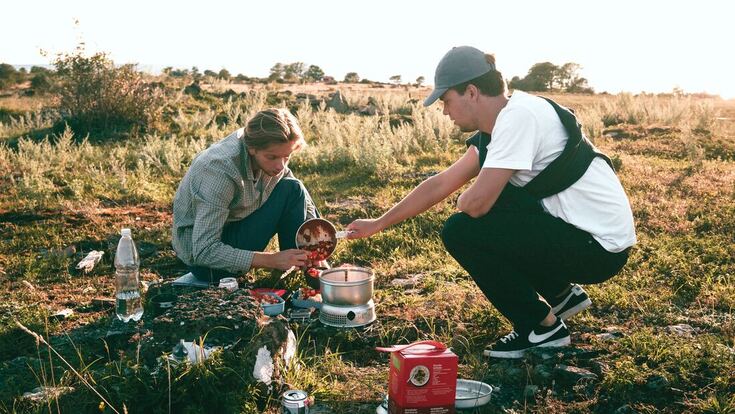 Two men listen and communicate while prepping food outdoors at a campsite.