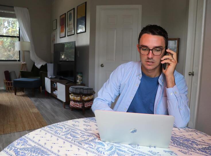 A man with glasses talks on his smartphone by an Apple laptop computer and asks questions about hearing aids.