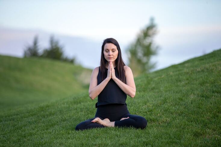 A woman wearing a black tank top and pants sits on a grassy, green field. She has tinnitus and meditates.
