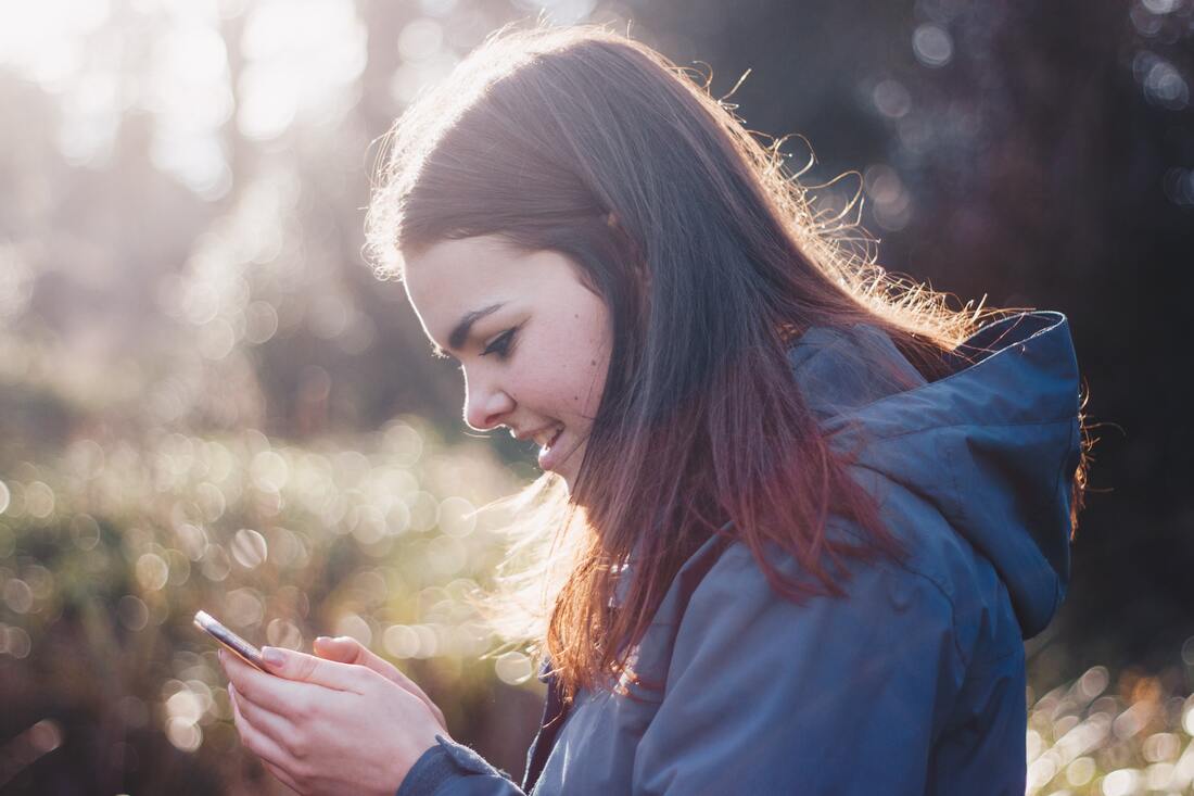 A girl in a hooded coat smiles and looks at her phone while using a caption app with her hearing aids.