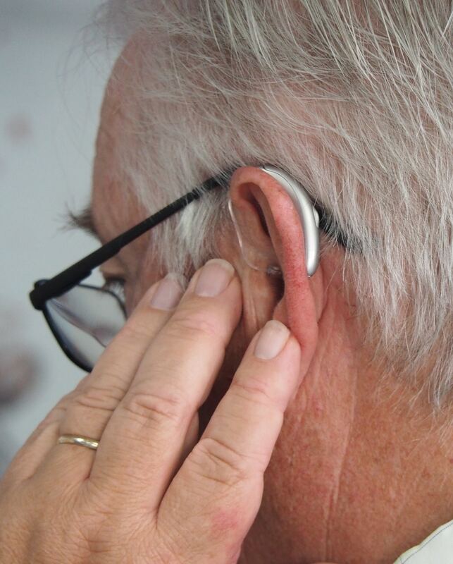 A man with white hair and black-framed eyeglasses places his hand on his ear with a silver behind-the-ear hearing aid in it.