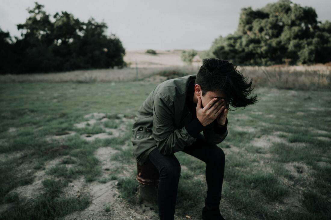 A man with tinnitus sits hunched over in a grassy area and places his hands over his face in frustration.