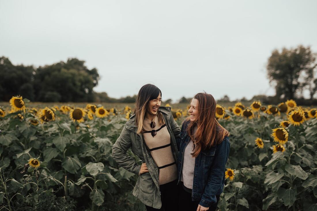 Two women laugh while standing in a field of tall, yellow sunflowers with green leaves and stalks.