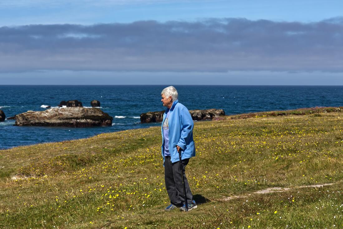woman-with-short-white-hair-wearing-periwinkle-blue-shirt-light-blue-jacket-black-pants-walks-in-the-green-grass-by-the-ocean